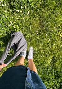 Veste grise tenue à la main, jupe en denim, chaussettes blanches et baskets grises sur l'herbe verte avec des marguerites blanches en arrière-plan.