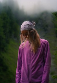Jonge vrouw met een witte bandana en paarse jas kijkt omlaag, omringd door een mistig bos met hoge groene bomen.