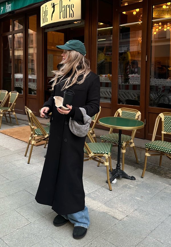 Schwarzer langer Mantel, hellblaue Jeans, grüner Hut, hält eine Kaffeetasse und eine beige Tasche. Der Hintergrund zeigt Caféstühle und einen grünen Tisch.