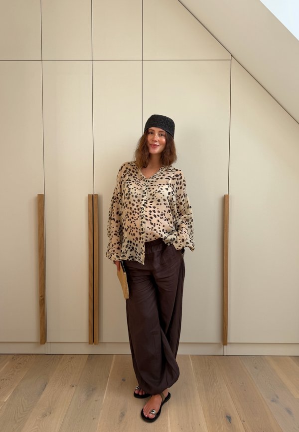 Femme debout à l’intérieur portant un chemisier imprimé léopard, un pantalon large foncé, un foulard noir sur la tête et des sandales, tenant une petite pochette beige.