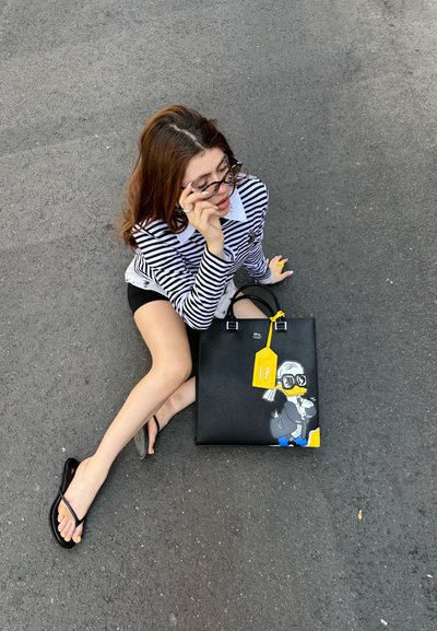 Joven mujer con camiseta de rayas y gafas sentada sobre el asfalto, sosteniendo un bolso negro con un pato de dibujos animados y una etiqueta amarilla.