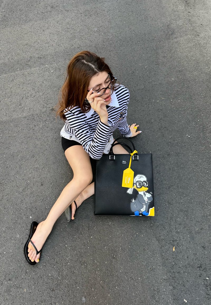 Jovem mulher a usar uma camisa às riscas e óculos, sentada no asfalto, segurando uma bolsa preta com um pato em cartoon e uma etiqueta amarela.
