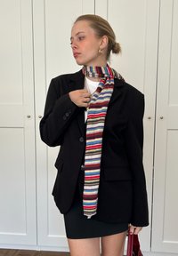 Black blazer featuring a double-button closure, paired with a multicoloured striped scarf. A short black skirt and red handbag complete the outfit.