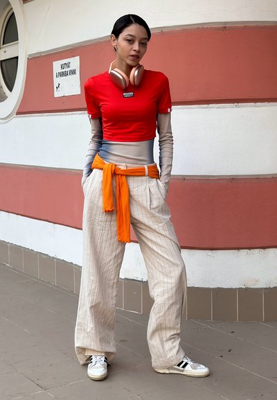 Top corto rojo de manga corta con detalles blancos, camiseta de manga larga gris claro debajo, pantalones de pierna ancha beige a rayas, y un cinturón naranja. Zapatillas blancas.