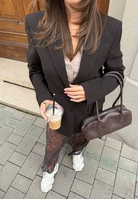 Čierny blazer na béžovej košeli na gombíky, kombinovaný s priesvitnými čipkovými nohavicami a bielymi teniskami. Drží ľadový nápoj a hnedú kabelku.