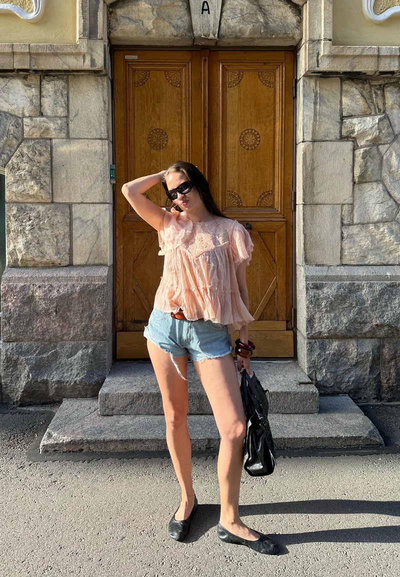 Femme en blouse rose et short en jean pose avec des lunettes de soleil et un sac noir devant une porte en bois ornée sur un bâtiment en pierre.