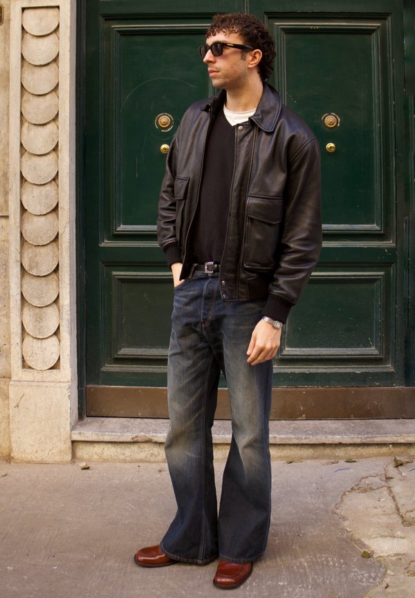 Hombre con chaqueta de cuero negra, suéter negro, vaqueros acampanados azules, zapatos marrones y gafas de sol, de pie junto a una puerta verde oscuro con pomos dorados.