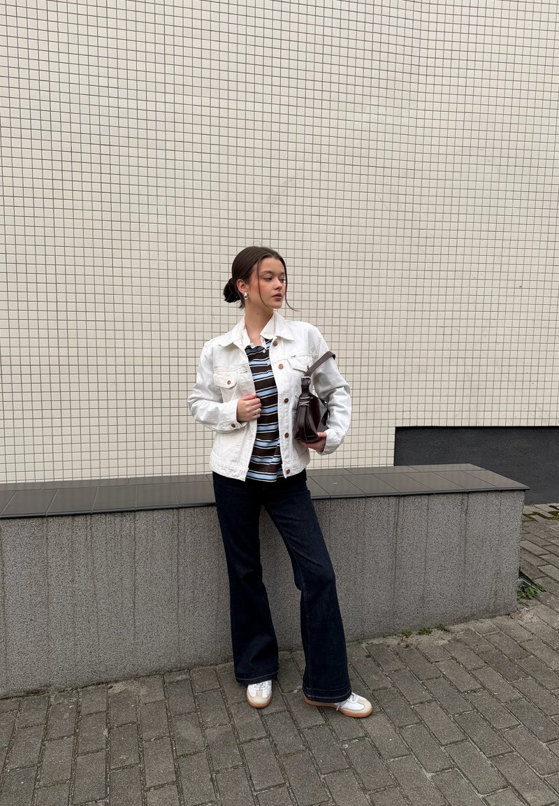 Fiatal nő fehér dzsekiben, csíkos ingben, sötét farmerban, fehér sportcipőben, barna táskát tartva, egy csempézett fal és egy szürke pad mellett áll kint.