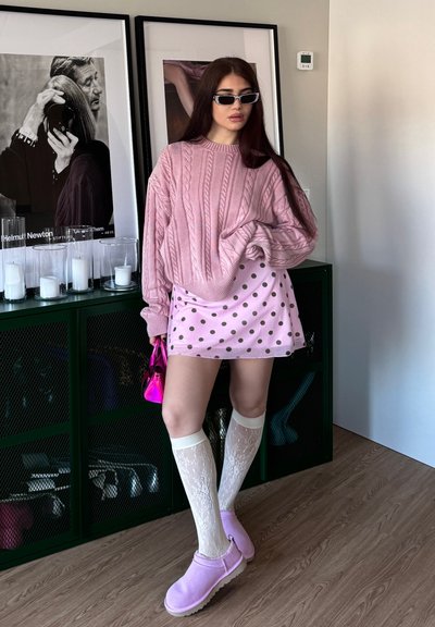 Mujer con un suéter rosa de punto grueso de gran tamaño, falda rosa con lunares, calcetines blancos de encaje hasta la rodilla y zapatos rosas sin cordones, sostiene una bolsa metálica rosa.