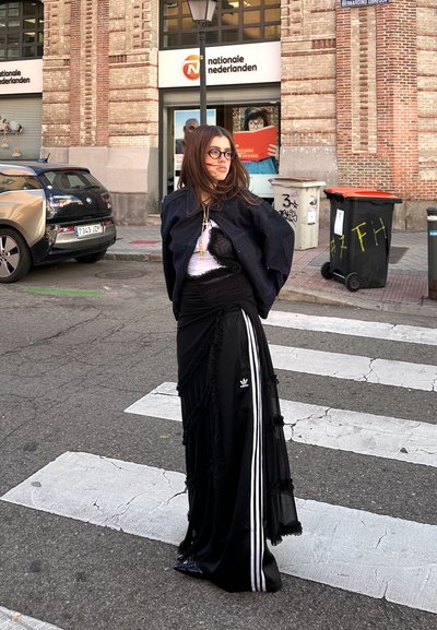 Falda negra con capas y detalles de flecos y rayas blancas, combinada con una chaqueta de mezclilla oscura y una blusa transparente. Los accesorios incluyen collares en capas.