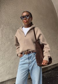 Γυναίκα που φοράει υπερμεγέθη γυαλιά ηλίου, μπεζ πουλόβερ με φερμουάρ, ανοιχτόχρωμα τζιν, καφέ ζώνη και κρατά μια μεγάλη καφέ τσάντα ώμου από σουέτ.