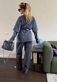 Femme aux longs cheveux blonds attachés en queue de cheval, portant des vêtements larges et sombres et des lunettes de soleil, tenant un sac à main gris, debout près d'une table avec des livres et une lampe bleue.