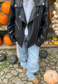Person i ljusblå jeans, bruna skor, grå hoodie och svart skinnjacka står bland orange och gröna pumpor på kullerstensbelagd mark.