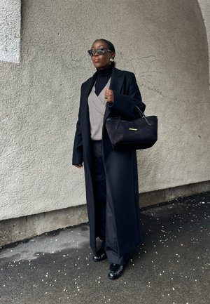 Mujer con abrigo largo negro, suéter beige sobre un cuello alto negro, pantalones negros, zapatos negros, gafas de sol, llevando bolso negro, de pie junto a una pared texturizada.