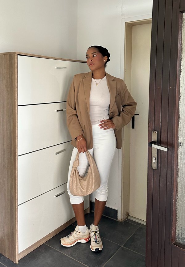 Beige blazer worn over a white top, paired with white capri pants, a beige handbag, and multi-coloured sneakers. Smooth textures, modern design, and functional elements.