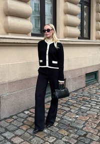 Γυναίκα που φοράει μαύρη και λευκή ζακέτα, μαύρο παντελόνι με φαρδιά μπατζάκια, γυαλιά ηλίου και κρατά μια μαύρη τσάντα, στέκεται σε λιθόστρωτο δρόμο.
