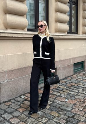 Mujer con cárdigan en blanco y negro, pantalones negros de pierna ancha, gafas de sol y bolso negro, de pie en una calle empedrada.