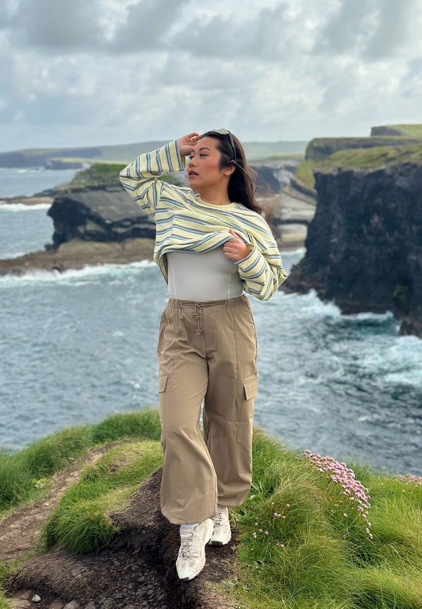 Jeune femme en pantalon beige et à manches longues rayées se tient sur une falaise herbeuse près de l'océan avec une côte rocheuse et un ciel nuageux en arrière-plan.