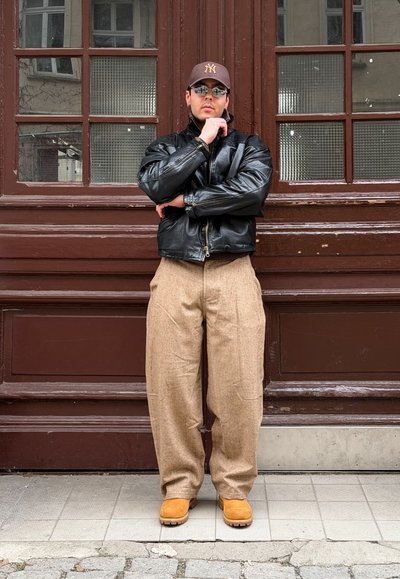 Hombre con chaqueta negra de cuero, pantalones anchos beige, botas marrones claras, gorra y gafas de sol, de pie con los brazos cruzados junto a una puerta de madera marrón con paneles de vidrio.