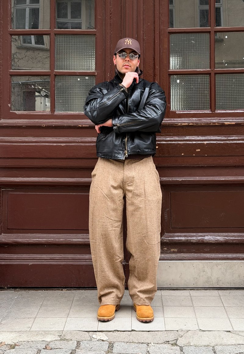 Mężczyzna w czarnej skórzanej kurtce, beżowych szerokich spodniach, brązowych butach, czapce i okularach przeciwsłonecznych stoi z założonymi rękami przy brązowych drewnianych drzwiach z przeszklonymi panelami.