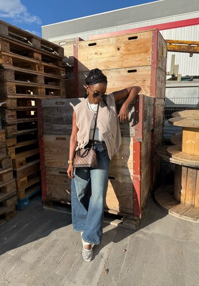 Chaleco sin mangas color beige claro, blusa blanca de canalé y jeans azules anchos. Bolso marrón con logo, mocasines blancos, gafas de sol, contra palets de madera.