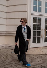 Manteau long noir sur un pull gris, chemise blanche et cravate bleu-vert; pantalon gris; portant des lunettes de soleil; tenant un sac noir; baskets bleues.