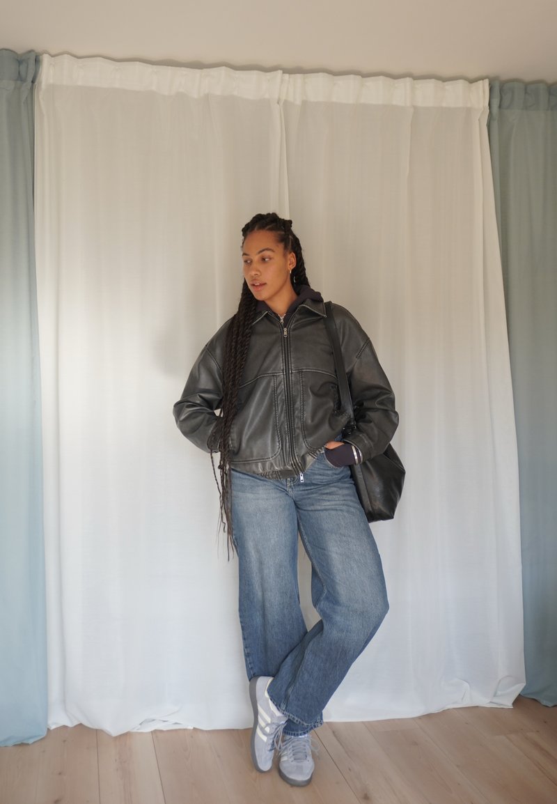 Giacca in pelle nera corta con zip, abbinata a jeans larghi blu e sneakers grigie. La modella tiene una borsa nera contro uno sfondo chiaro.