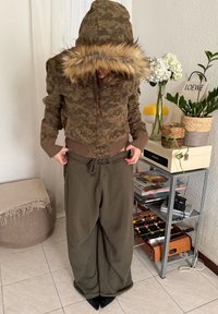 Personne portant une veste camouflage marron avec capuche en fourrure et un pantalon vert ample, debout à l'intérieur près d'une table d'appoint avec des plantes et des livres.