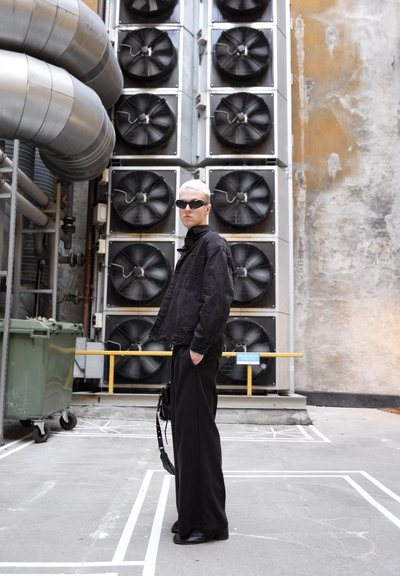 Chaqueta de mezclilla negra, pantalones de pierna ancha negros, gafas de sol negras de gran tamaño. Fondo industrial con grandes ventiladores de refrigeración y superficies grises.