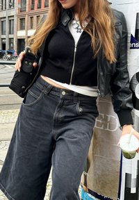 Μαύρο κοντό πουλόβερ με φερμουάρ, ψηλόμεσες σκούρες τζιν culottes και μια μαύρη μίνι τσάντα. Κρατάει ένα ποτό με καλαμάκι.