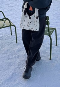 Personne debout sur la neige portant des bottes et un pantalon noirs, tenant un sac à main blanc à taches noires à côté de deux chaises métalliques vertes.