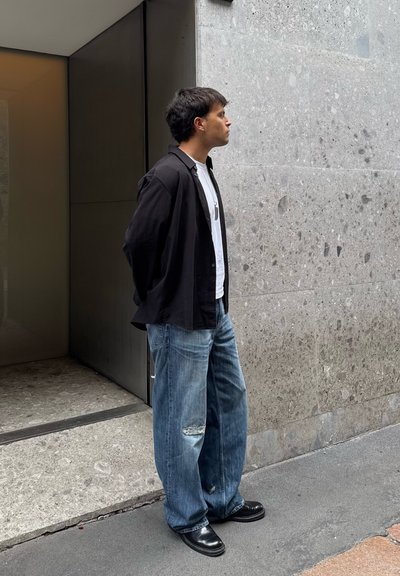Una persona con una camisa negra abotonada, camiseta blanca y pantalones vaqueros azules sueltos, está de pie frente a una pared gris texturizada con una puerta cerca.