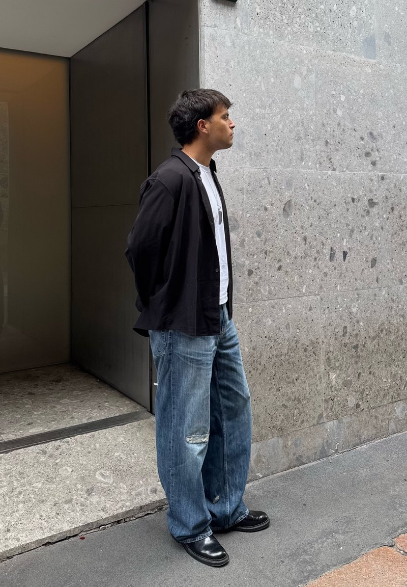 Cilvēks ar melnu ar pogām aiztaisāmu kreklu, baltu t-kreklu un vaļīgām zilām džinsu biksēm stāv pie teksturētas pelēkas sienas netālu no durvīm.