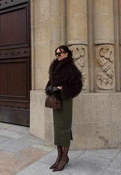 Abrigo de piel de pelo largo marrón, falda lápiz de cuero verde oliva, bolso marrón, tacones altos marrones, guantes negros, gafas de sol oscuras, fondo de piedra texturizada.