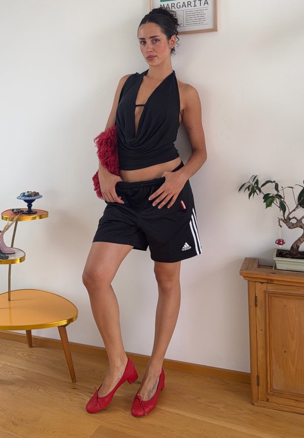 Kvinde i sort haltertop og Adidas-shorts læner sig op ad væggen, holder en rød håndtaske, iført røde hæle, ved siden af et træskab og et gult bord.