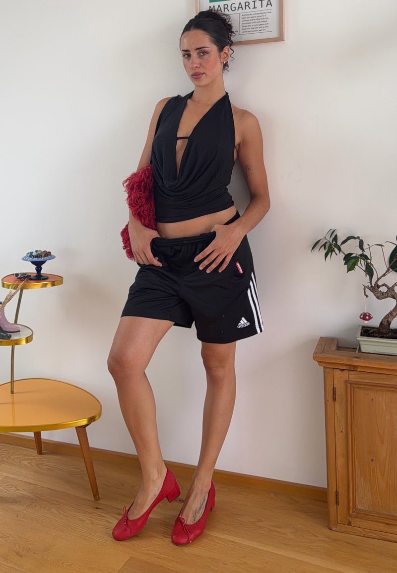 Donna con top nero a halter e pantaloncini Adidas si appoggia al muro, tiene in mano una borsa rossa, indossa tacchi rossi, accanto a un armadio di legno e un tavolo giallo.