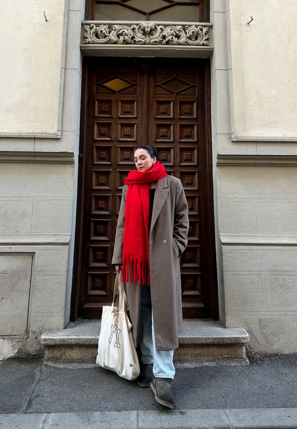 Καφέ μάλλινο παλτό, κόκκινο φραγκόσκαφο κασκόλ, γκρι παντελόνι και καφέ μπότες. Κρατάει μια κρέμα τσάντα ώμου με λογότυπο. Ξύλινη πόρτα με γεωμετ�ρικό μοτίβο στο φόντο.