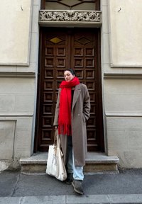 Καφέ μάλλινο παλτό, κόκκινο φραγκόσκαφο κασκόλ, γκρι παντελόνι και καφέ μπότες. Κρατάει μια κρέμα τσάντα ώμου με λογότυπο. Ξύλινη πόρτα με γεωμετρικό μοτίβο στο φόντο.