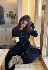 Ung kvinne i svart antrekk som sitter med bena i kors på flislagt gulv ved vinduet, holder en kopp kaffe, med telefon og veske i nærheten.