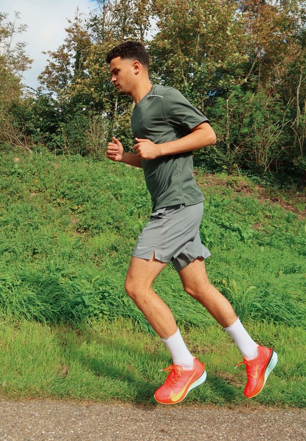Un hombre con una camiseta verde, pantalones cortos grises y zapatillas de correr de color naranja intenso trota por un sendero junto a árboles frondosos.