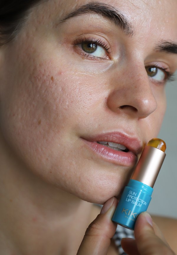Gros plan d'une personne tenant un baume à lèvres de protection solaire bleu et or près de ses lèvres, montrant la texture naturelle de la peau et des yeux verts.