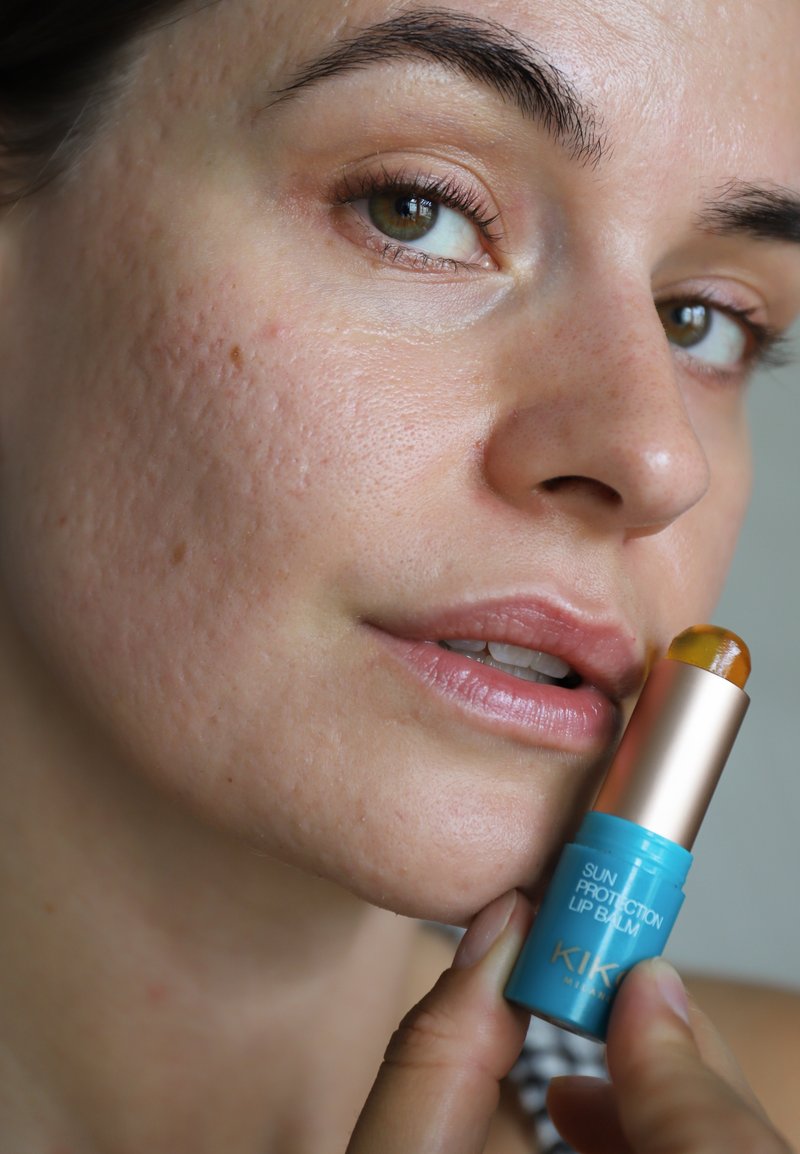 Primer plano de una persona sosteniendo un bálsamo labial de protección solar azul y dorado cerca de sus labios, mostrando la textura natural de la piel y ojos verdes.
