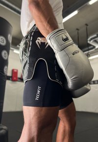 Melnas sporta šortus ar sietu dizainu un logo, apvienojot ar pelēkām boksa cimdiem, kuru virsma ir ar teksturētu saķeri. Fona attēlā redzami treniņu rīki.