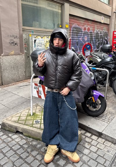 Persona con chaqueta de plumas negra, vaqueros holgados y botas beige, de pie en una calle adoquinada cerca de scooters estacionados y paredes cubiertas de grafitis.