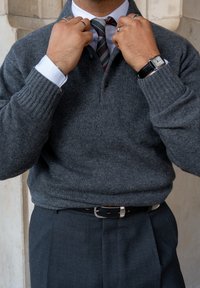 Pullover grigio a maglia con polsini a costine, indossato sopra una camicia bianca e una cravatta a righe. Pantaloni grigio scuro e una cintura nera con fibbia argentata.
