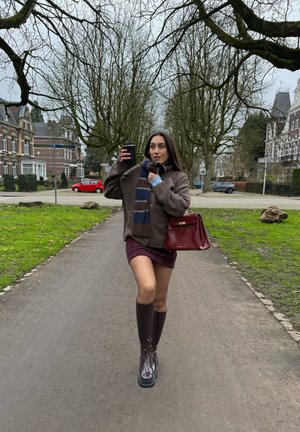 Joven mujer caminando por un sendero bordeado de árboles, sosteniendo una taza negra, vistiendo un suéter marrón, bufanda a rayas, falda burdeos, botas y llevando un bolso rojo.