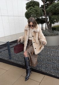 Cappotto in pelliccia ecologica beige con grandi bottoni, abbinato a un maglione beige e una gonna a quadri. Stivali neri con tacco e una borsa bordeaux completano il look.