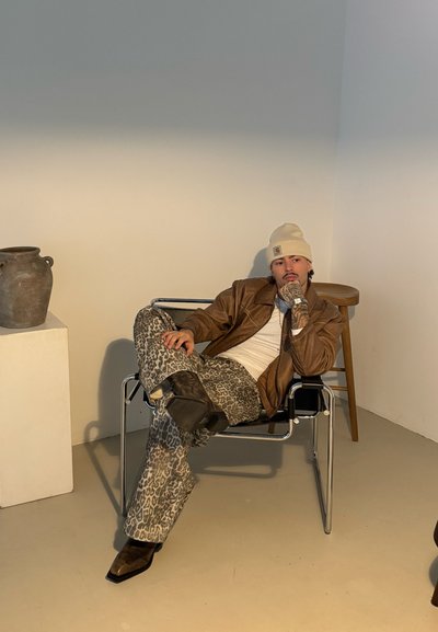 Chaqueta de cuero marrón, camisa blanca, pantalones de leopardo, botas negras. Sentado en una silla metálica con una maceta gris y un taburete de madera cerca.
