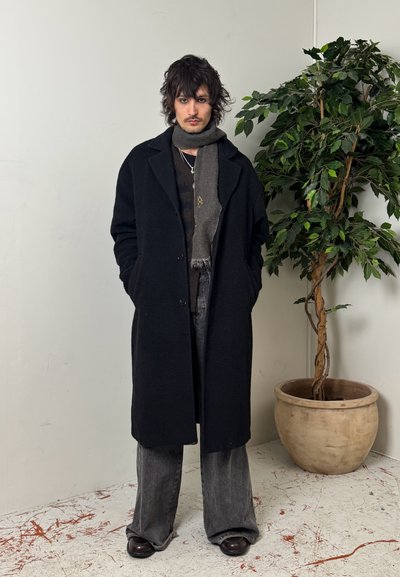 Hombre con cabello despeinado que lleva un abrigo largo negro, una bufanda gris, pantalones anchos grises y zapatos oscuros, de pie en el interior junto a una planta grande en maceta.