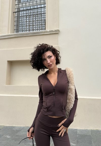 Mujer de cabello rizado y oscuro, vestida con un top marrón ajustado con cremallera y pantalones, llevando una chaqueta peluda beige sobre un hombro, de pie en una acera.